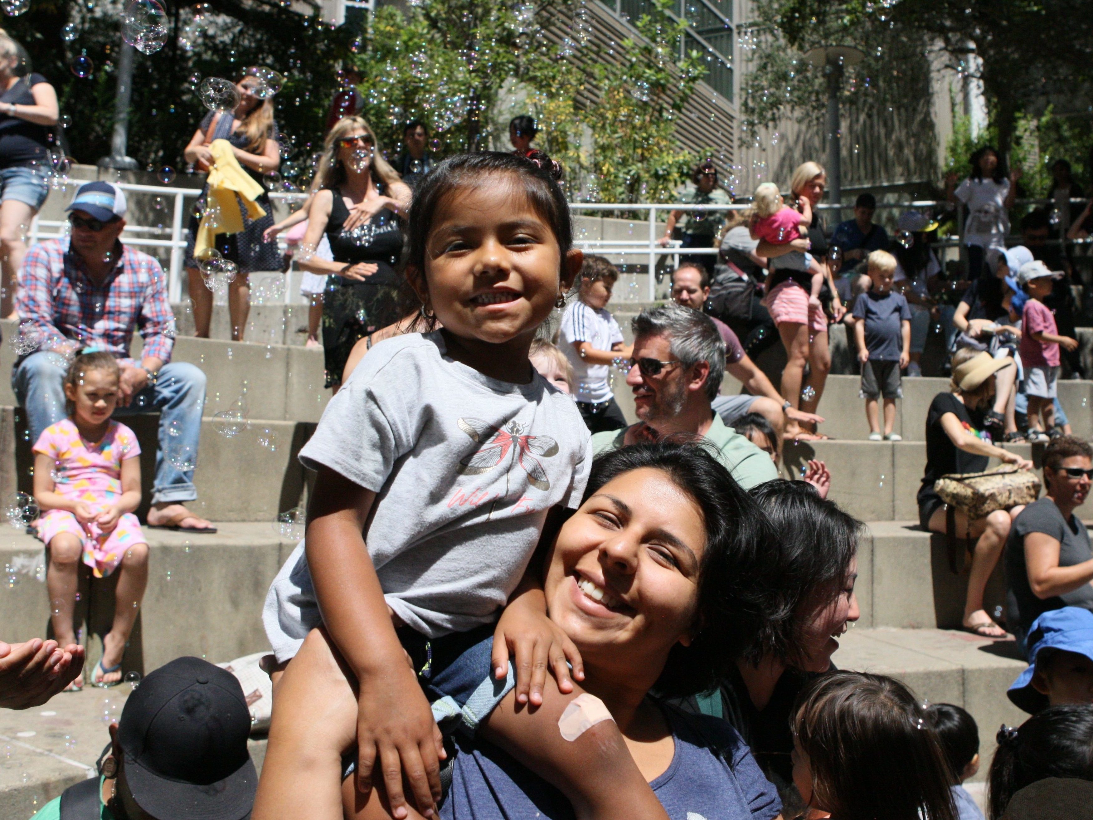 Smiling visitors in the courtyard during a sunny day, with a background filled with other visitors and  bubbles. 